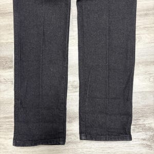 May include: A pair of dark charcoal grey denim trousers featuring a straight-leg cut with visible vertical creases running down the centre of each leg. The fabric has a textured weave and a consistent dark tone throughout the garment's visible length.