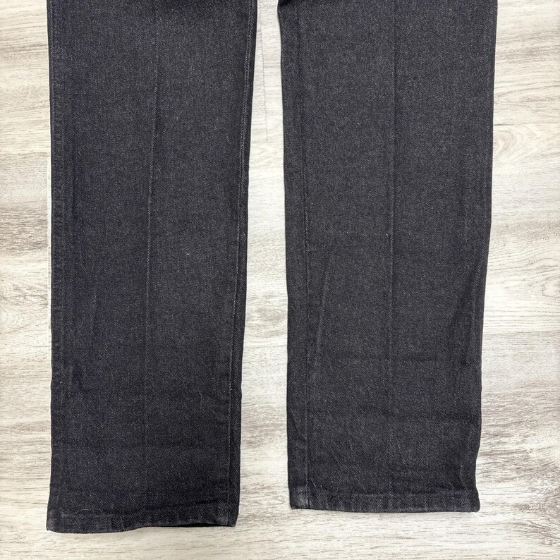 May include: A pair of dark charcoal grey denim trousers featuring a straight-leg cut with visible vertical creases running down the centre of each leg. The fabric has a textured weave and a consistent dark tone throughout the garment's visible length.