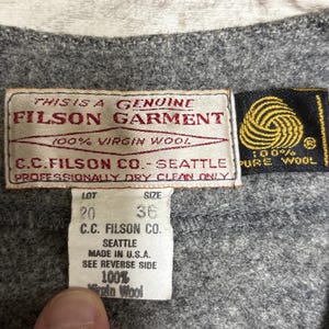 May include: Close-up of a gray garment with multiple tags. The main tag reads "FILSON GARMENT" and "100% Virgin Wool." A smaller tag indicates size 36. The garment is made by C.C. Filson Co. in Seattle, USA. The garment is made of 100% pure wool.