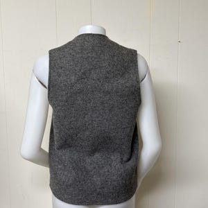 May include: A gray vest made of a textured fabric. The vest is sleeveless with a rounded neckline. The back of the vest is visible, displayed on a white mannequin. The vest is a solid color and appears to be a medium weight.
