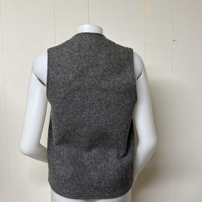 May include: A gray vest made of a textured fabric. The vest is sleeveless with a rounded neckline. The back of the vest is visible, displayed on a white mannequin. The vest is a solid color and appears to be a medium weight.