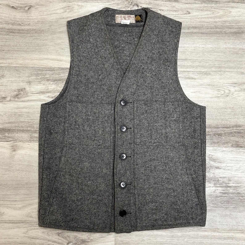 May include: A gray wool vest with a V-neck and a button-up front. The vest has two front pockets and a small label near the collar. The vest is made of a thick, textured fabric.