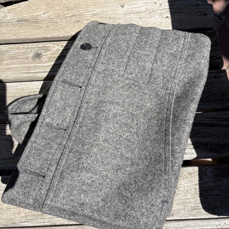 May include: A gray wool coat with a button closure and a notched collar. The coat has a classic design with vertical seams and a tailored fit. The coat is folded on a wooden surface.
