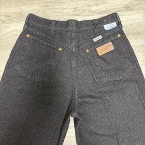 May include: A pair of black denim jeans featuring a high-rise waist, two back pockets with gold-tone rivets, and a tan leather patch with the Wrangler logo. A white tag is attached to the waistband, displaying the text Size 13 and Wrangler.