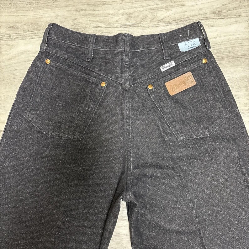 May include: A pair of black denim jeans featuring a high-rise waist, two back pockets with gold-tone rivets, and a tan leather patch with the Wrangler logo. A white tag is attached to the waistband, displaying the text Size 13 and Wrangler.