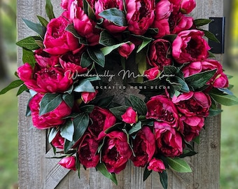 May include: A pink peony wreath with green leaves. The wreath is hanging on a wooden door. The text "Wonderfully Made Home Decor" is visible on the wreath.