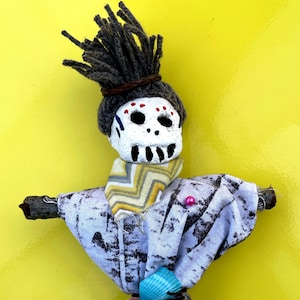 May include: A handmade doll with a skull-like face, yarn hair, and a patterned scarf. The doll's body is made of fabric and decorated with seashells and a pink bead. The doll has a rustic, folk art aesthetic.