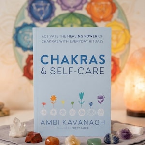 Peut inclure: Un livre bleu clair intitulé Chakras & Self-Care par Ambi Kavanagh avec un sous-titre sur le pouvoir de guérison et les rituels quotidiens. La couverture présente des illustrations de fleurs colorées et des symboles de chakras blancs.