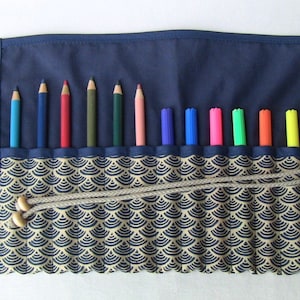 Può includere: Un rotolo per matite in tessuto blu con un motivo geometrico beige e blu. Il rotolo ha 13 fessure per matite e pennarelli. Il rotolo è legato con una corda beige.