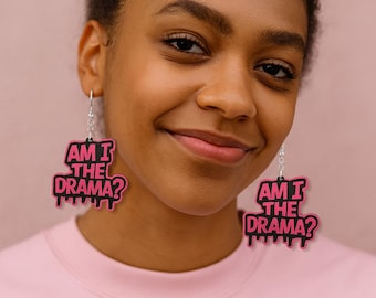 Pode incluir: Brincos pendurados rosa e pretos com o texto "AM I THE DRAMA?". O texto está em negrito. Os brincos penduram-se de ganchos prateados. O fundo é rosa liso.