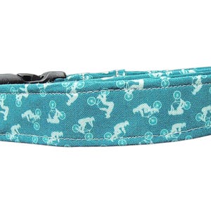 Op de afbeelding: Teal hondenriem met een herhalend wit patroon van fietsstuntrijders. De halsband heeft een zwarte plastic sluiting. De stof is een stevige katoenmix.