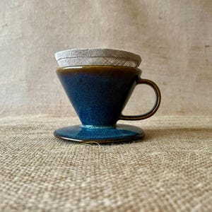 Op de afbeelding: Een keramische koffiefilter met een diepblauwe glazuurlaag, een bruine rand en één handvat. De kegelvormige houder staat op een ronde voet en bevat een lichtbeige stoffen filterinzetstuk dat in de bovenste opening is geplaatst.