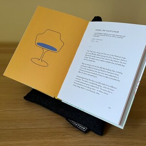 Op de afbeelding: Een open boek getiteld Model 150 Tulip Chair op een zwarte stoffen standaard. De linkerpagina heeft een gele achtergrond met een zwarte lijntekening van een stoel met een blauwe zitting. De rechterpagina bevat beschrijvende tekst over de stoel.