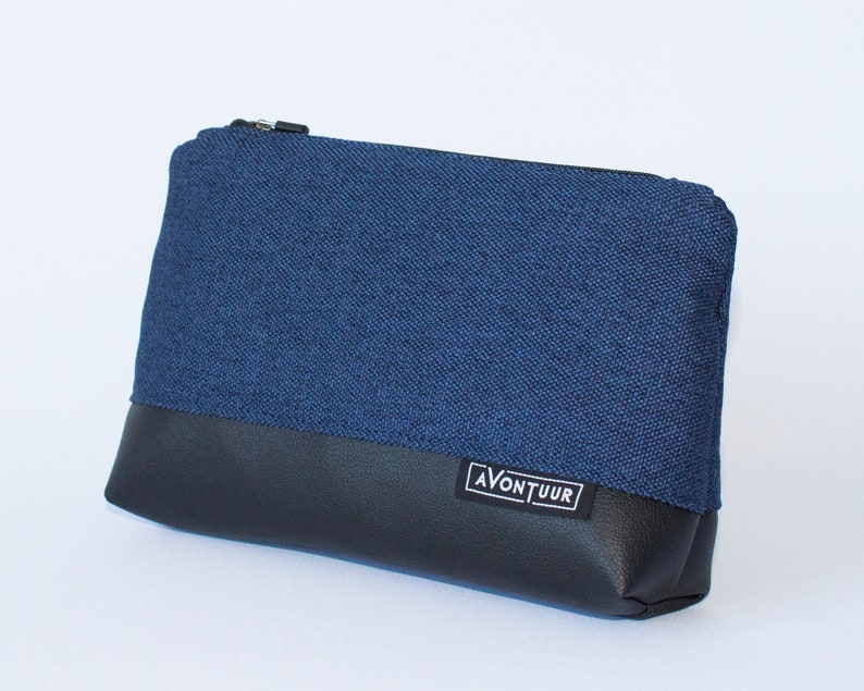 May include: A rectangular pouch with a navy blue fabric upper and a black leather-like bottom. The pouch has a black zipper closure and a small white label with the word "AVONTUUR" in black. The pouch is designed for carrying small items.