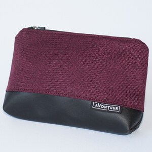May include: A rectangular pouch with a burgundy fabric upper and a black leather-like lower section. The pouch has a silver zipper and a small white label with the text "AVONTUUR". The pouch is likely used for carrying small items.