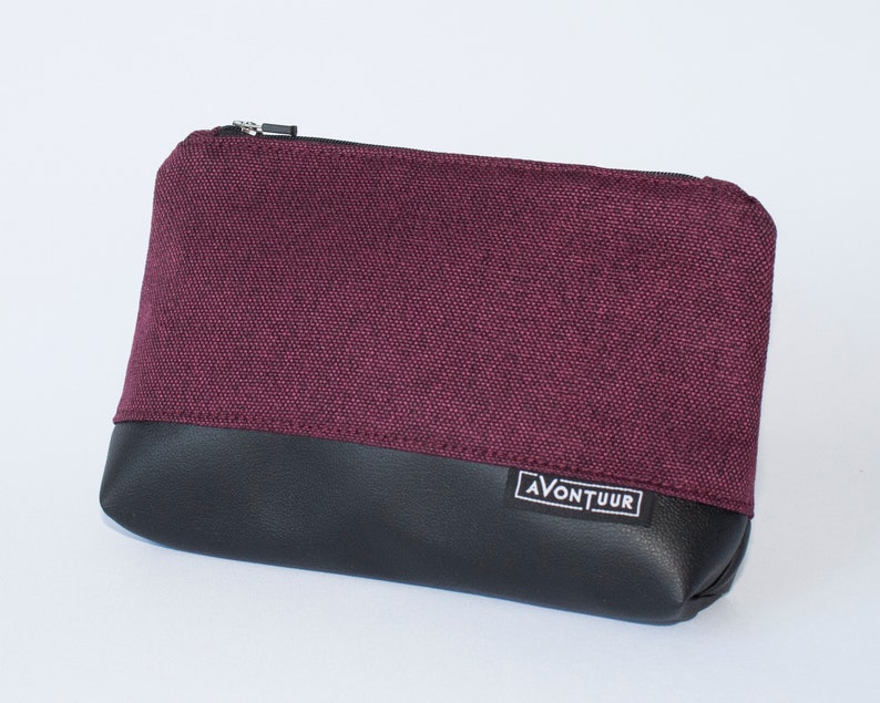 May include: A rectangular pouch with a burgundy fabric upper and a black leather-like lower section. The pouch has a silver zipper and a small white label with the text "AVONTUUR". The pouch is likely used for carrying small items.