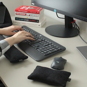 Peut inclure: Des mains tapent sur un clavier noir avec une souris noire et un repose-poignets noir. Il y a deux repose-poignets noirs sur le bureau. Une pile de livres se trouve sur le bureau derrière le clavier. Un écran d'ordinateur noir est sur le bureau derrière le clavier.