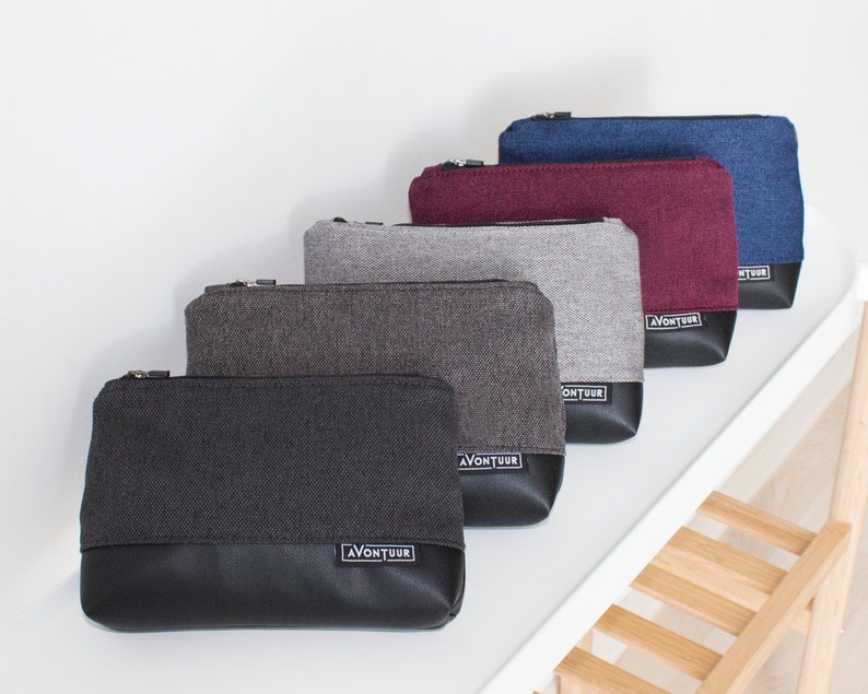 May include: A collection of five rectangular zippered pouches in various colors. The pouches feature a textured fabric upper and a black leather-like bottom. Each pouch has a small label that reads "AVONTUUR". Colors include black, gray, burgundy, and blue.