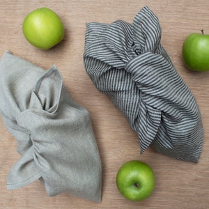 May include: Two fabric wraps tied in a knot, one in a solid light gray and the other in a black and white striped pattern. Three bright green apples are positioned around the fabric bundles, which are folded to create a secure, knotted closure.