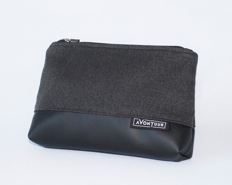 May include: A rectangular, two-toned pouch with a zipper closure. The top half is a dark gray woven fabric, and the bottom half is black faux leather. The label "AVONTUUR" is visible on the lower right.