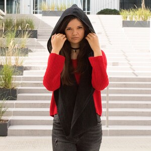May include: A woman wearing a black hooded sweater over a red sweater. She is standing in front of a white staircase.