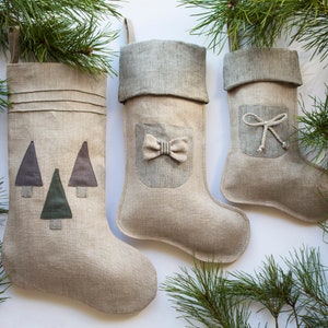 Peut inclure: Trois chaussettes de Noël en lin beige avec des boucles de suspension. La chaussette de gauche présente trois arbres en tissu gris et vert. Celle du milieu a un revers et une poche à rayures grises avec un petit nœud. Celle de droite a un revers et une poche à rayures grises avec un lien en corde blanche.