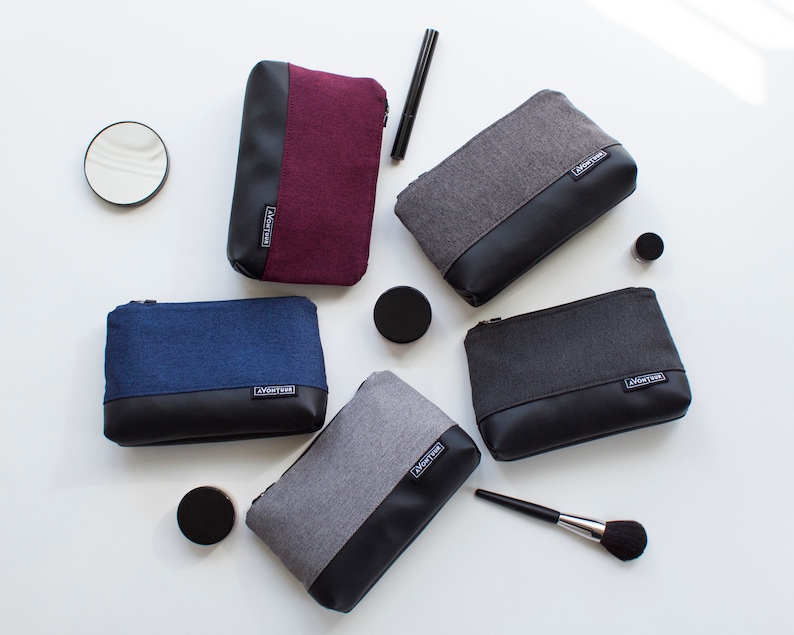May include: Five zippered cosmetic bags in various colors: burgundy, gray, navy blue, and charcoal. Each bag has a fabric upper and a black leather-look lower section. The bags are arranged with makeup accessories, including a mirror and a brush.