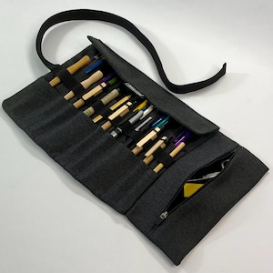 May include: A dark gray fabric pencil roll with a long strap, featuring multiple elastic slots holding various pens, pencils, and tools, along with a zippered pocket at the bottom containing small items and a small black label with white text.