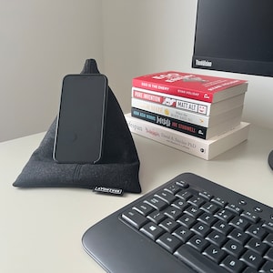 May include: A black phone stand with a black phone resting on it. The stand is on a white desk with a black keyboard and a stack of books. The books have titles such as "SEO is the Enemy", "Pure Invention", "How Asia Works", and "When the Body Says No".