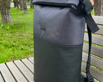 May include: A roll-top backpack featuring a dark gray textured fabric upper section and a smooth black base. The bag includes a black plastic side-release buckle, a top carry handle, adjustable shoulder straps, and a small rectangular brand label.