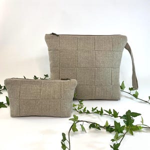 May include: Two beige fabric pouches with a quilted design. The larger pouch is rectangular with a handle, and the smaller one is a rectangular cosmetic bag. Both have a zipper closure. Green vines with leaves are in the foreground.
