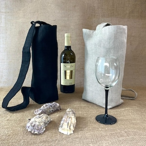Könnte beinhalten: Eine schwarze Weinflaschentasche aus Stoff mit langem Trageriemen, eine beigefarbene Weinflaschentasche, eine Glasweinflasche mit weißem Etikett mit der Aufschrift Soave, ein Klarglas mit schwarzem Stiel und drei rohe Austernschalen.