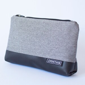 May include: A rectangular gray and black zippered pouch. The top portion is gray fabric, and the bottom is black faux leather. A small white label with the word "AVONTUUR" is on the lower front. The zipper is black.