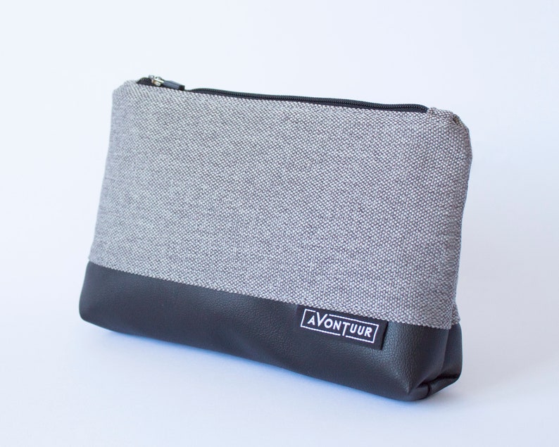 May include: A rectangular gray and black zippered pouch. The top portion is gray fabric, and the bottom is black faux leather. A small white label with the word "AVONTUUR" is on the lower front. The zipper is black.