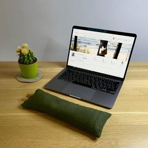 Könnte beinhalten: Eine dunkelgrüne, rechteckige Handgelenkauflage aus Stoff vor einem grauen Laptop, der einen Online-Shop anzeigt. Links neben dem Laptop steht ein kleiner grüner Kaktus in einem gerippten, lindgrünen Topf auf einem runden Untersetzer.