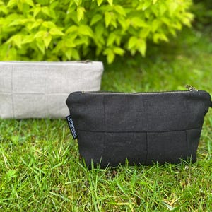 Peut inclure: Deux pochettes rectangulaires à fermeture éclair, une noire et une beige clair, présentant un motif quadrillé matelassé. Chaque pochette possède une petite étiquette en tissu noir avec le texte AVONTUUR sur le côté et une dragonne.