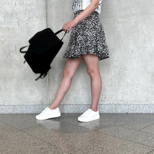 Peut inclure: Une jupe noire à motif floral blanc avec un ourlet à volants, associée à un t-shirt blanc, des baskets blanches et un sac à dos noir. La jupe est portée à la taille et tombe au-dessus des genoux, présentant un design simple et fonctionnel.