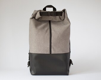Könnte beinhalten: Ein Rolltop-Rucksack mit einem melierten grauen Oberteil und einem einfarbigen schwarzen Boden. Der Rucksack verfügt über einen vertikalen schwarzen Reißverschluss in der Mitte, einen schwarzen Tragegriff und einen verstellbaren Schnallenriemen.