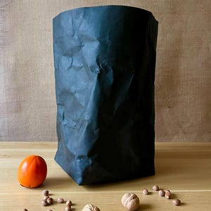 Könnte beinhalten: Ein schwarzer Papierbeutel steht auf einer Holzoberfläche, neben einer orangefarbenen Kaki, Walnüssen und Haselnüssen. Der Beutel hat eine zerknitterte Textur. Der Hintergrund ist ein neutraler Beigeton.
