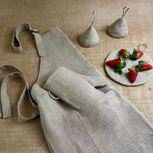 Op de afbeelding: Een grijze linnen schort met een grote zak en een bijpassende ovenwant. Het schort heeft verstelbare banden en een lus om op te hangen. Er zijn ook twee grijze linnen ovenwanten.