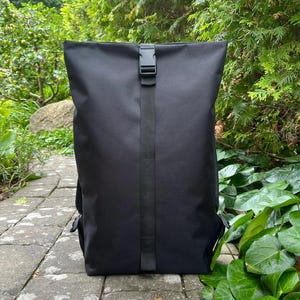 Op de afbeelding: Een effen zwarte roll-top rugzak met een verticale nylon band in het midden en een zwarte kunststof gespsluiting aan de bovenkant. De tas heeft een gladde, matte afwerking en een rechthoekige vorm met afgeronde hoeken aan de bovenzijde.