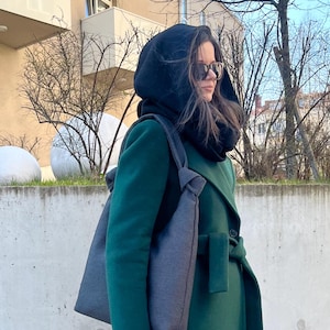 May include: A dark green coat with a matching hood and a gray tote bag. The coat has a belted waist and a long, flowing design. The tote bag is a simple, rectangular shape with a shoulder strap. The person is wearing sunglasses.