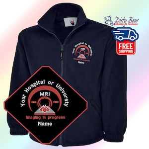 K&ouml;nnte beinhalten: Marineblaue Fleecejacke mit Rei&szlig;verschluss und gesticktem MRI-Design. Die Jacke hat einen hohen Kragen und ein kleines Logo auf der Brust. Das Design enth&auml;lt den Text "Your Hospital or University", "MRI", "Imaging in progress" und "Name".