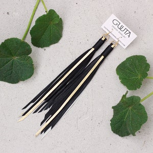 Può includere: Orecchini in pelle nera con dettagli dorati e design a frangia. Gli orecchini sono appesi a una superficie bianca con foglie verdi sullo sfondo. Il testo "GWTA DESIGN JEWELLERY" è su un cartoncino bianco.