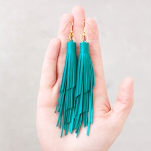 Op de afbeelding: Een paar turquoise blauwe kwast oorbellen met gouden haken. De oorbellen hangen aan een hand.