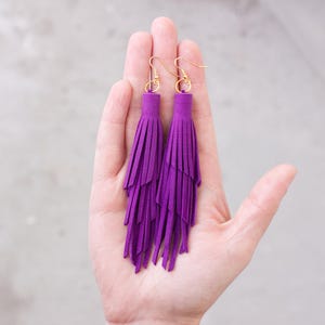 May include: A pair of vibrant purple tassel earrings with gold-colored hooks. The earrings are held in a hand, showcasing their layered design and rich color. The tassels are made of a soft material, creating a flowing, textured look.