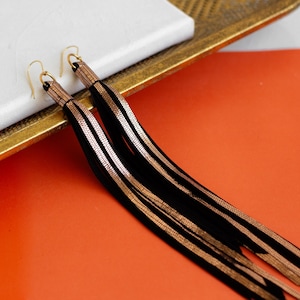 以下が含まれることがあります： 黒と金の長いタッセルイヤリングのペア。イヤリングはスエードと金属製で、フリンジのデザインです。