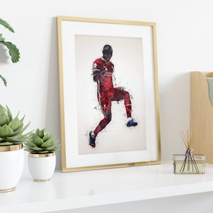 May include: A framed print of a soccer player in a red and white uniform, with a white background and a splash of color effect. The player is in mid-air, kicking the ball.