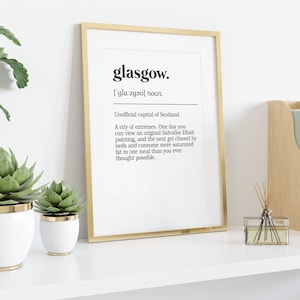 Peut inclure: Une impression encadr&eacute;e dor&eacute;e avec le mot "glasgow" en gras en noir et une d&eacute;finition du mot. L'impression est sur une &eacute;tag&egrave;re blanche avec un pot blanc avec une plante verte, un pot blanc plus petit avec une plante verte, un porte-revues en bois avec des magazines verts et un diffuseur en verre transparent avec des roseaux bruns.