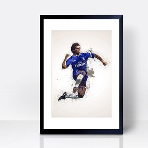 May include: A framed print of a soccer player in a blue jersey with white accents, running with the ball. The player is in mid-air, kicking the ball with his right foot. The background is a white splatter effect.